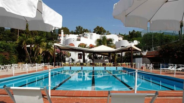 En swimmingpool med hvide liggestole og parasoller, omgivet af en belagt terrasse og en bygning med hvide vægge og orange døre.