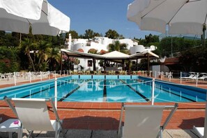 Una piscina con sillas de descanso blancas y sombrillas, rodeada por un patio de ladrillo y un edificio con paredes blancas y puertas naranjas.