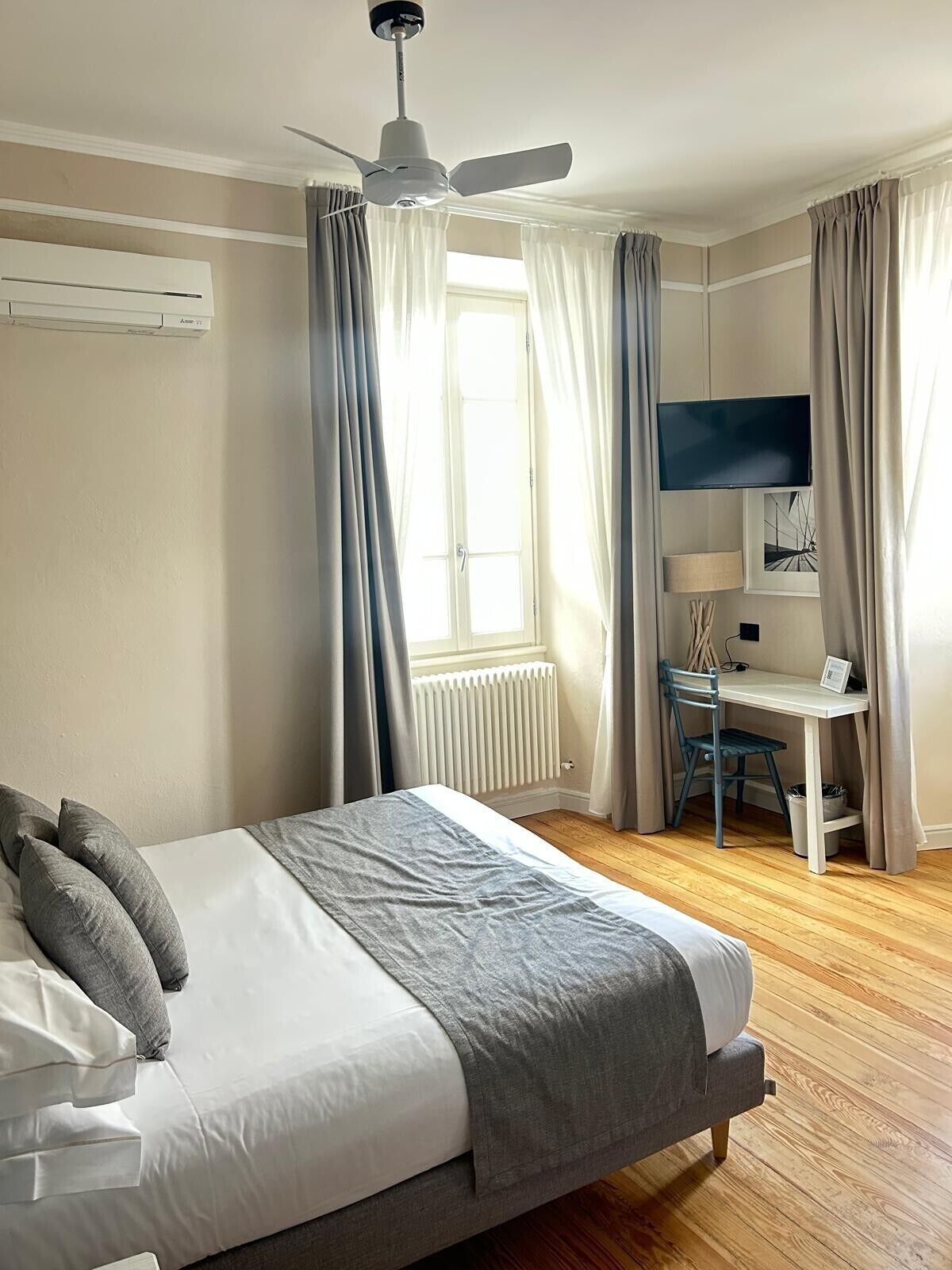 A bedroom with a bed, a desk, a chair, a television, a ceiling fan, and a window with curtains.