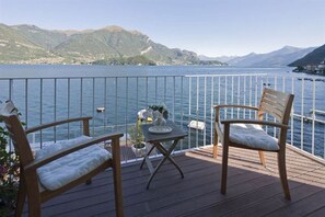 A deck with two wooden chairs and a small table, overlooking a lake with mountains in the background.