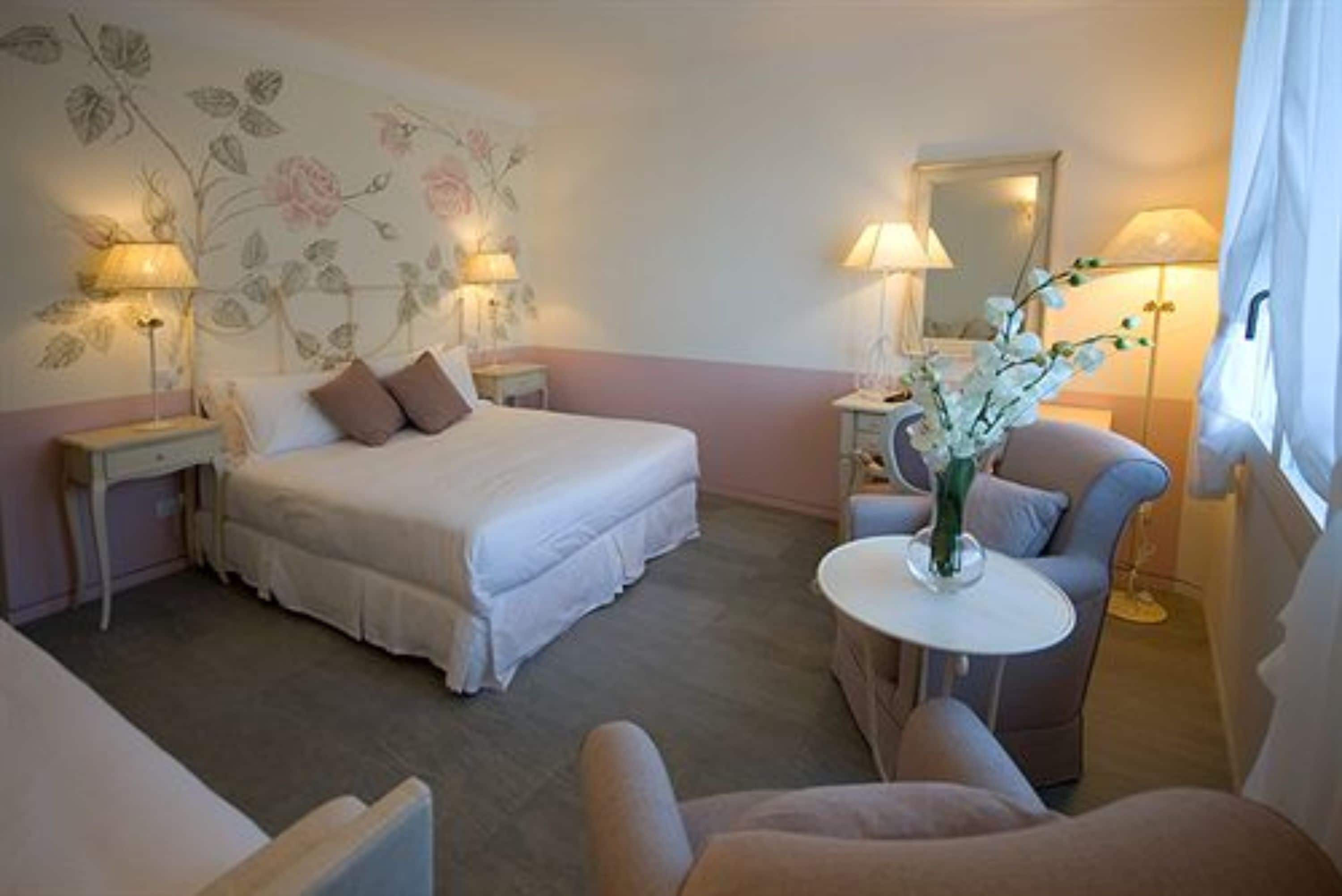 Una habitación de hotel con cama, mesitas de noche, una silla, una mesita pequeña y un jarrón con flores.