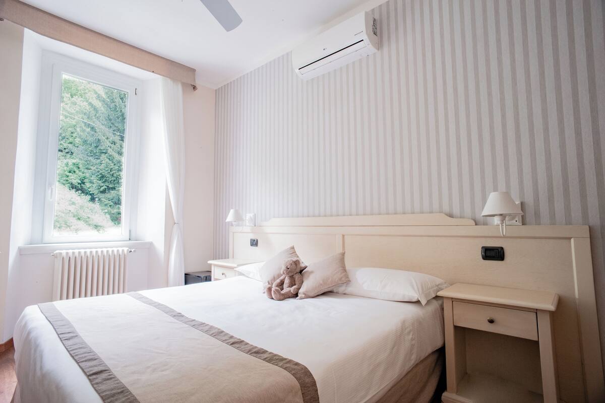 A neatly made bed with a beige headboard, a bedside table with a lamp, and a window with curtains.