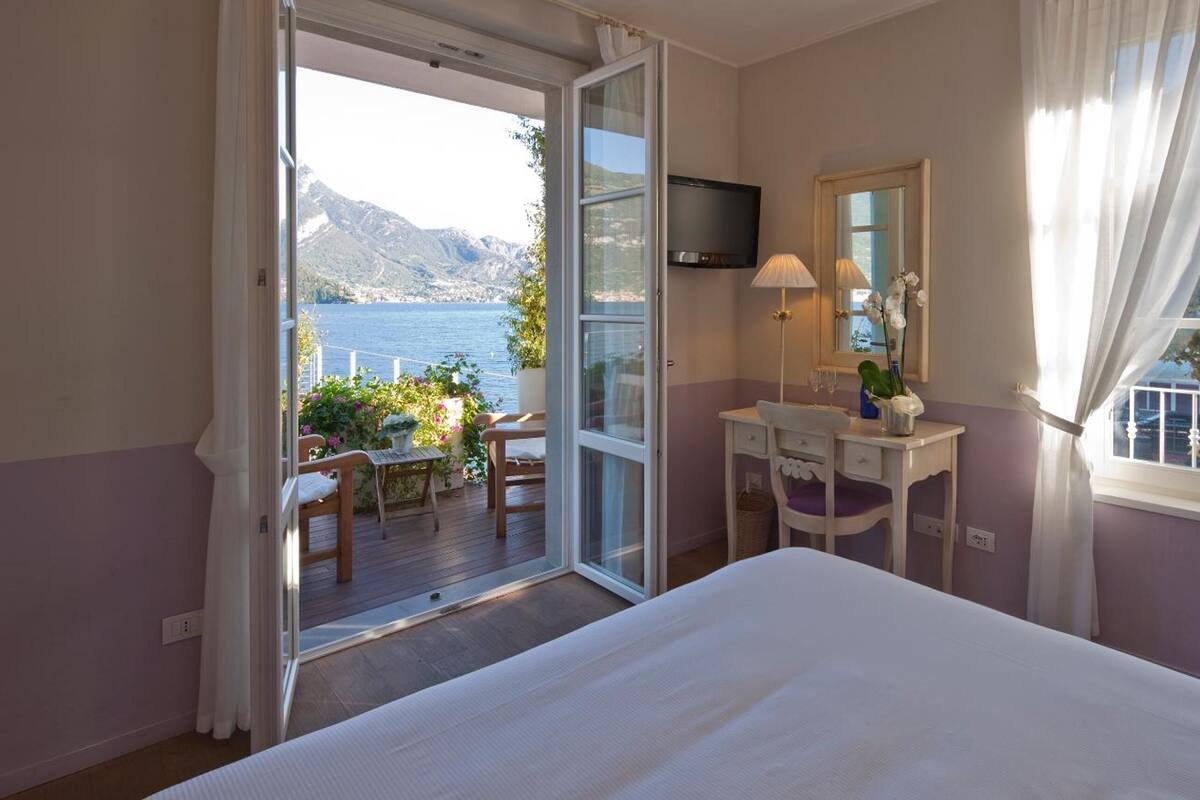 A bedroom with a bed, a desk, a TV, and a view of a lake and mountains.
