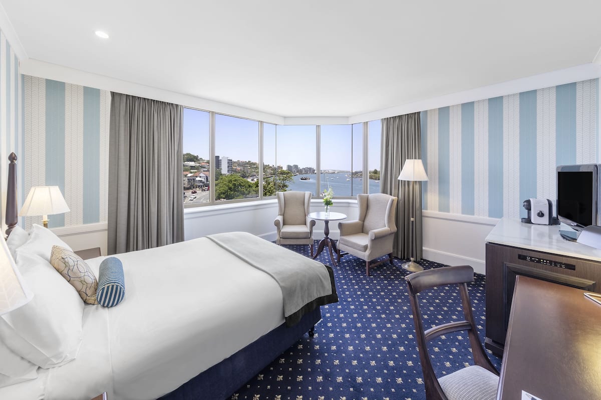 A hotel room with a large bed, two armchairs, a small round table, a desk with a TV, and a view of the city and water.