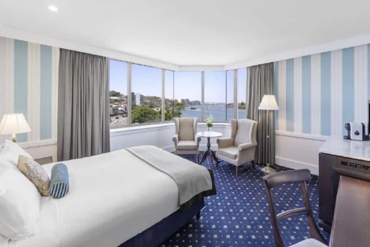 A hotel room with a bed, two chairs, a small table, a desk, and large windows offering a city view.