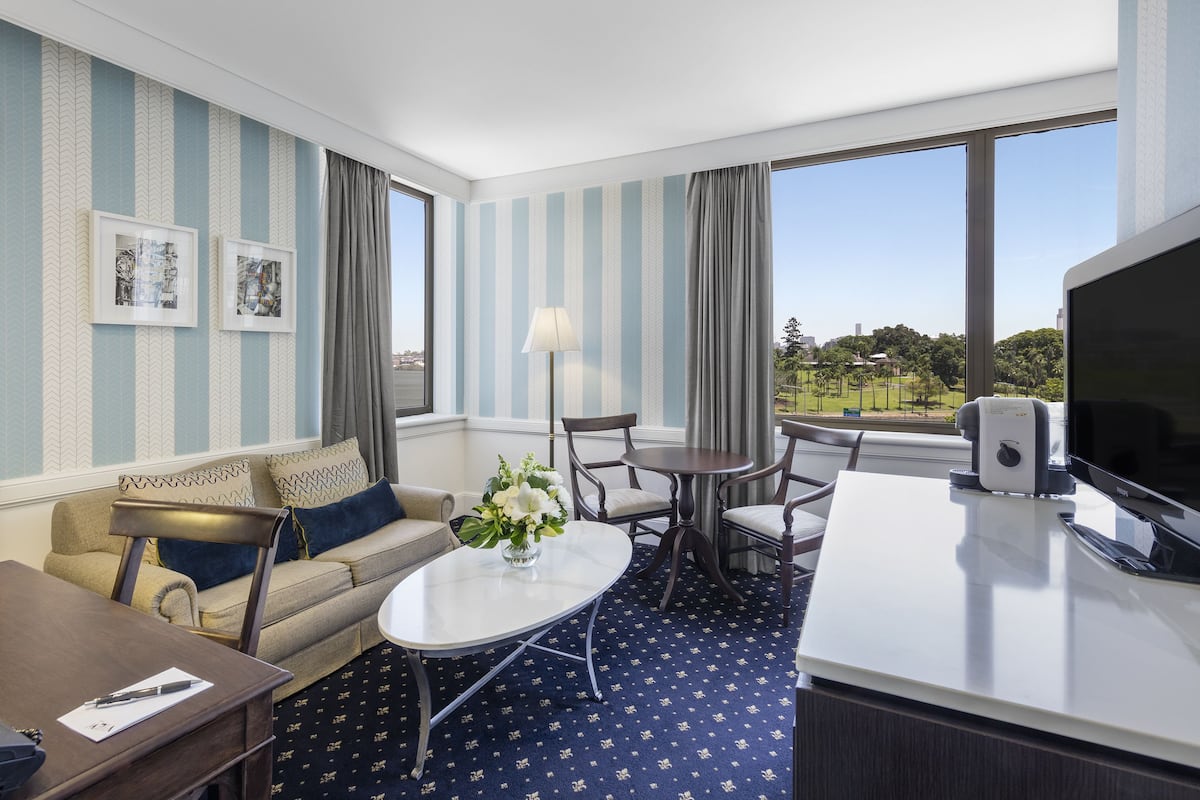A hotel room with a sofa, a dining table, a coffee table, a TV, and a large window offering a view of a park.