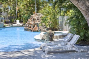 Piscina con sillas de descanso, rodeada de rocas y palmeras.