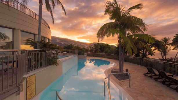 Une piscine avec vue sur le coucher de soleil, des palmiers et un bâtiment moderne.