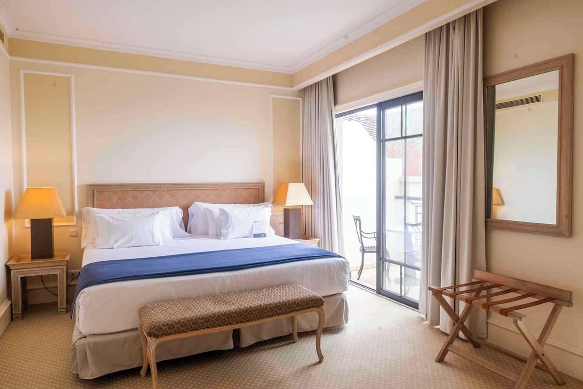 Une chambre d’hôtel avec un grand lit, deux lampes de chevet, un banc, une chaise en bois et une vue sur l’extérieur par une grande fenêtre.