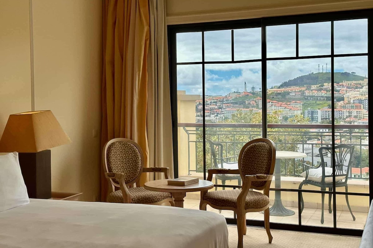 Habitación de hotel con cama, dos sillas, una mesita y vista a un paisaje urbano a través de ventanas amplias.