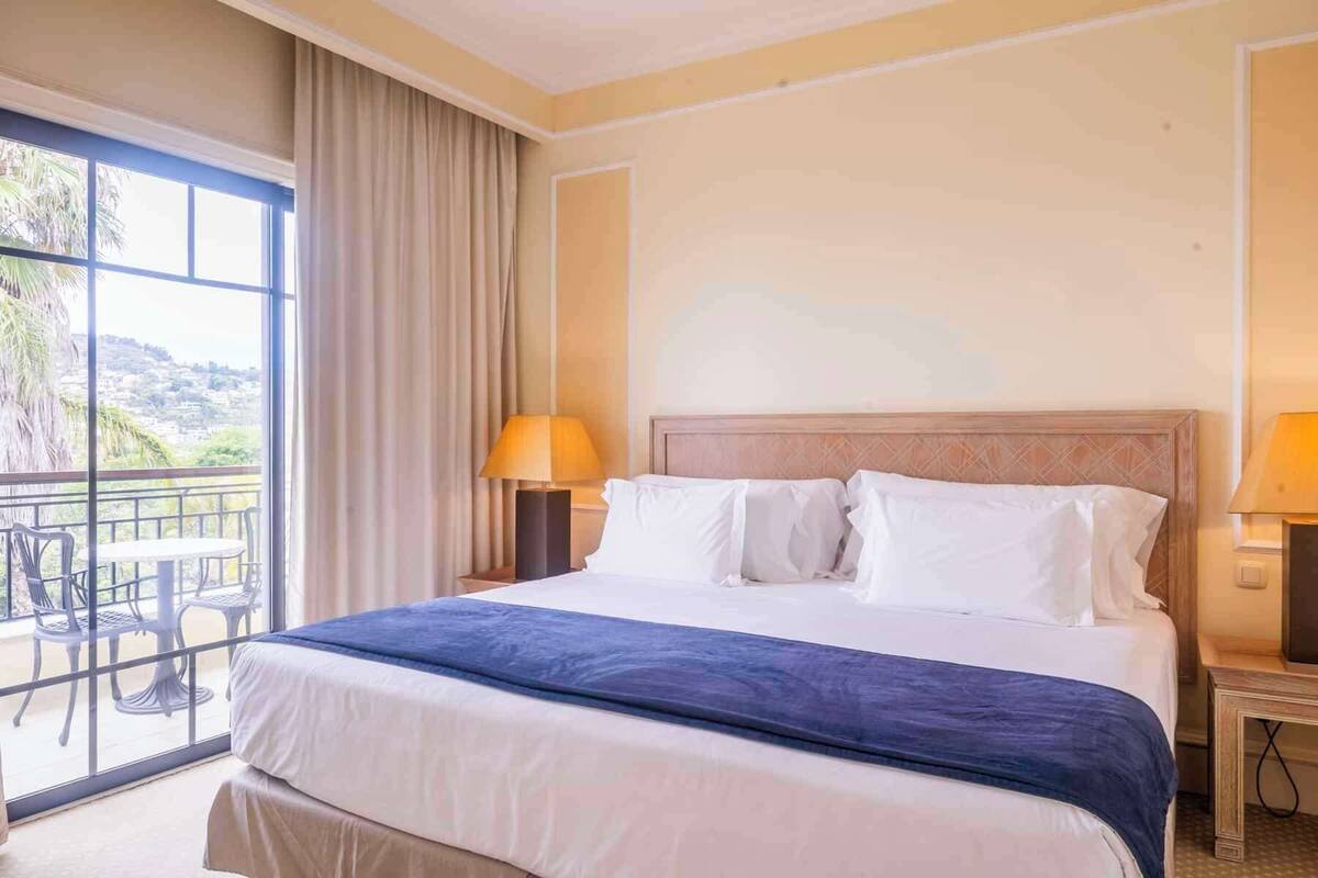Une chambre d’hôtel avec un grand lit, deux lampes de chevet, un balcon avec des chaises et une vue sur le paysage.