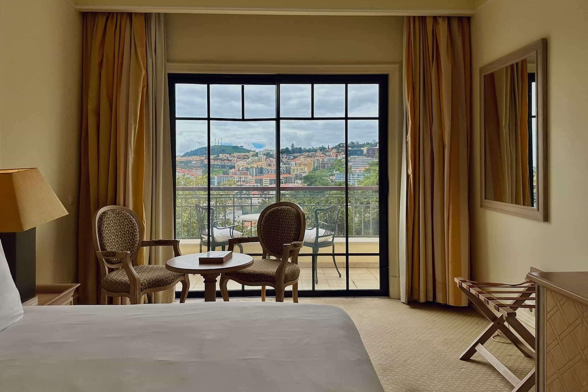 Ein Hotelzimmer mit einem großen Bett, zwei Stühlen, einem kleinen Tisch und Blick auf eine Stadtlandschaft durch ein großes Fenster.