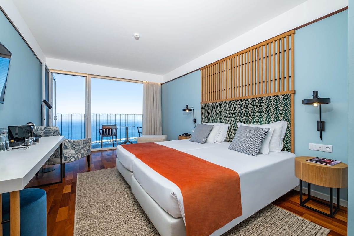 A modern hotel room with a large bed, a desk with a computer, a balcony with a view, and a patterned headboard.
