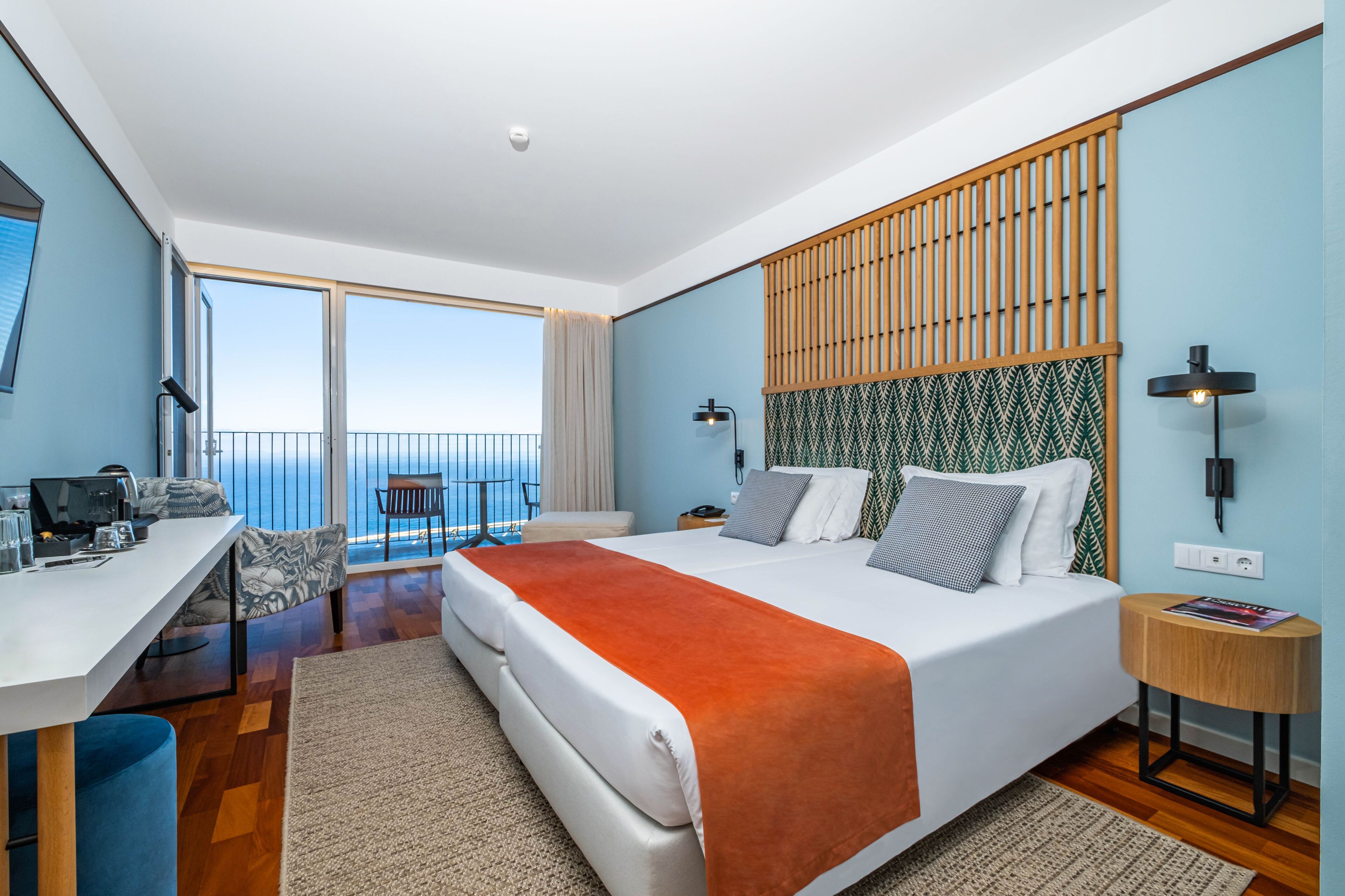 A modern hotel room with a large bed, a desk with a computer, a balcony with a view, and a patterned headboard.