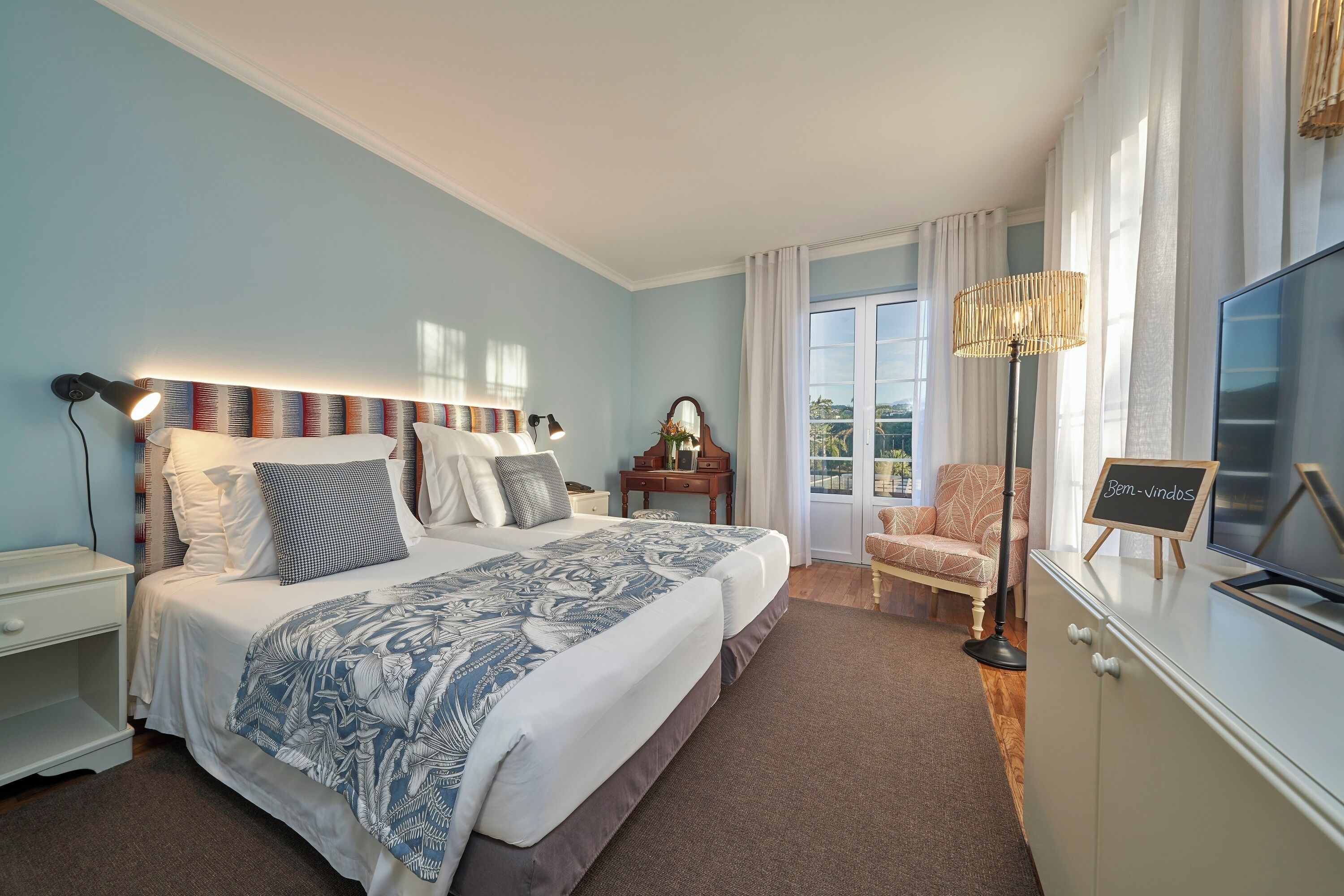 A bedroom with a bed, bedside table, lamp, chair, TV, and a view of the outdoors.