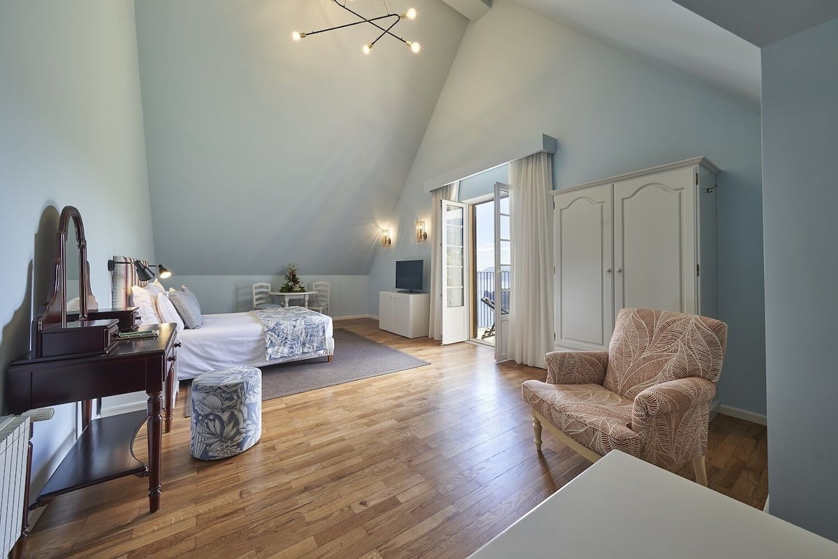 A spacious room with a sloped ceiling, a bed, a desk with a lamp, a chair, and a wardrobe.