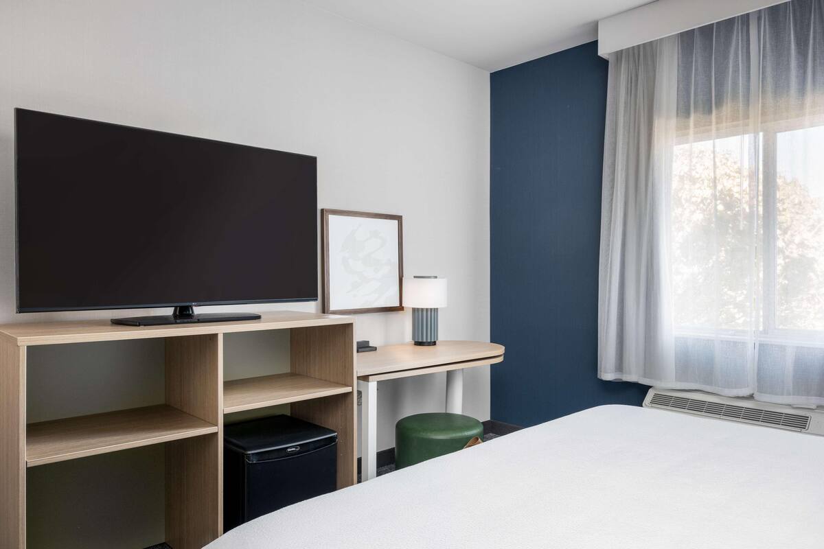 A hotel room with a bed, a flat-screen TV, a desk with a lamp, a waste bin, and a window with sheer curtains.