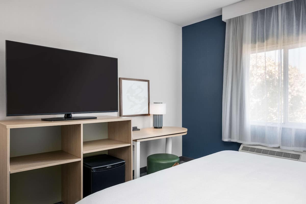 A hotel room with a bed, a flat-screen TV, a desk with a lamp, a waste bin, and a window with sheer curtains.
