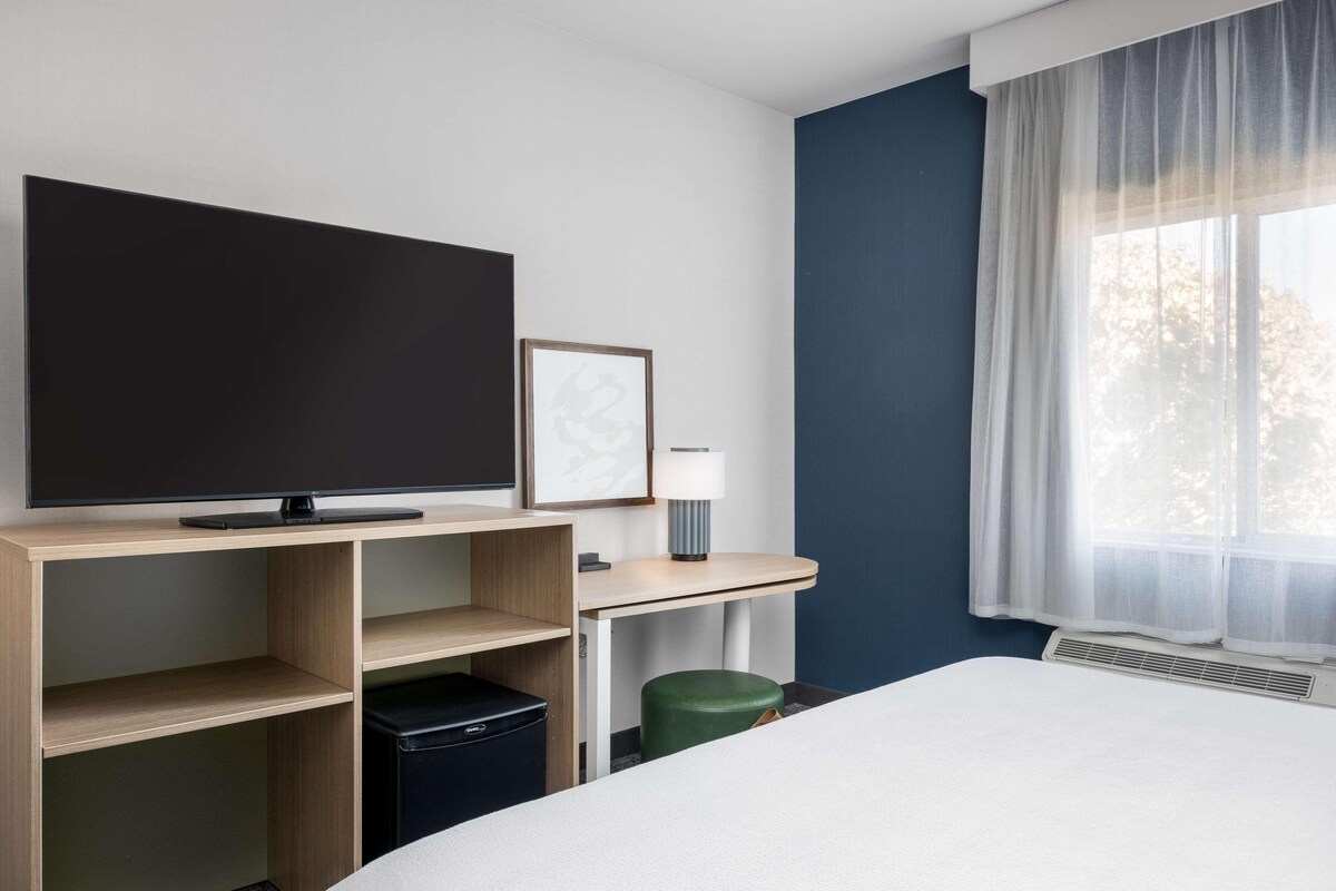 A hotel room with a bed, a flat-screen TV, a desk with a lamp, a waste bin, and a window with sheer curtains.