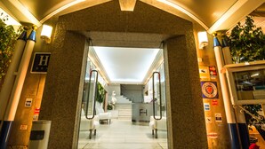 Entrada de hotel con mostrador de recepción, una escalera y un letrero con un símbolo 'H'.