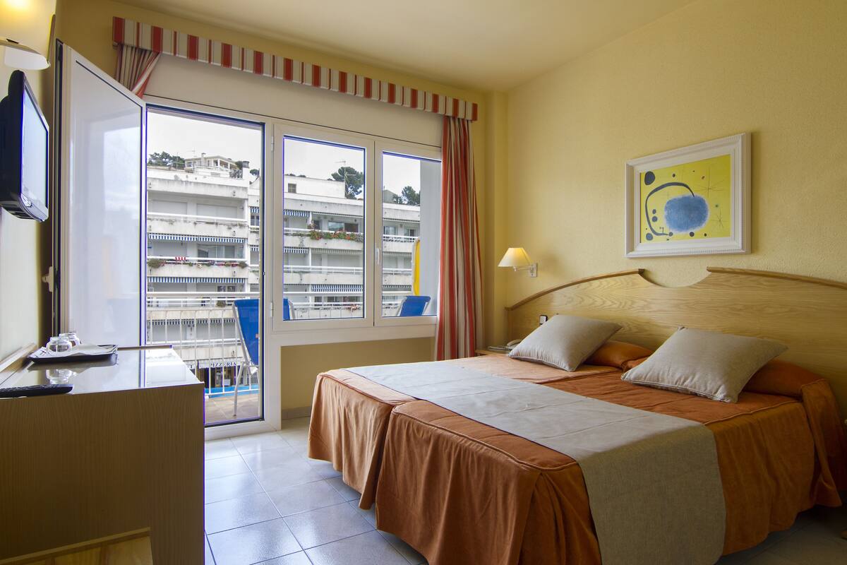 A hotel room with a bed, a desk, a chair, a window with a view of buildings, and a painting on the wall.