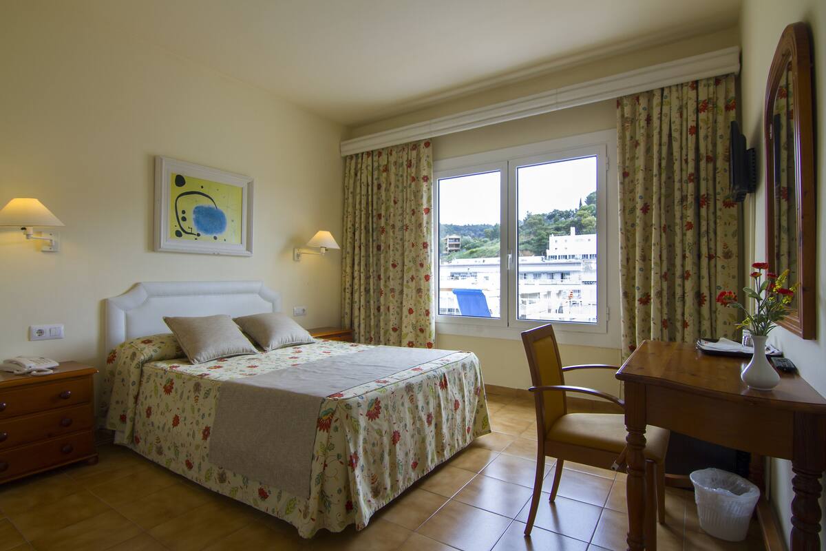 A hotel room with a bed, a desk, a chair, a window with curtains, a lamp, and a painting on the wall.
