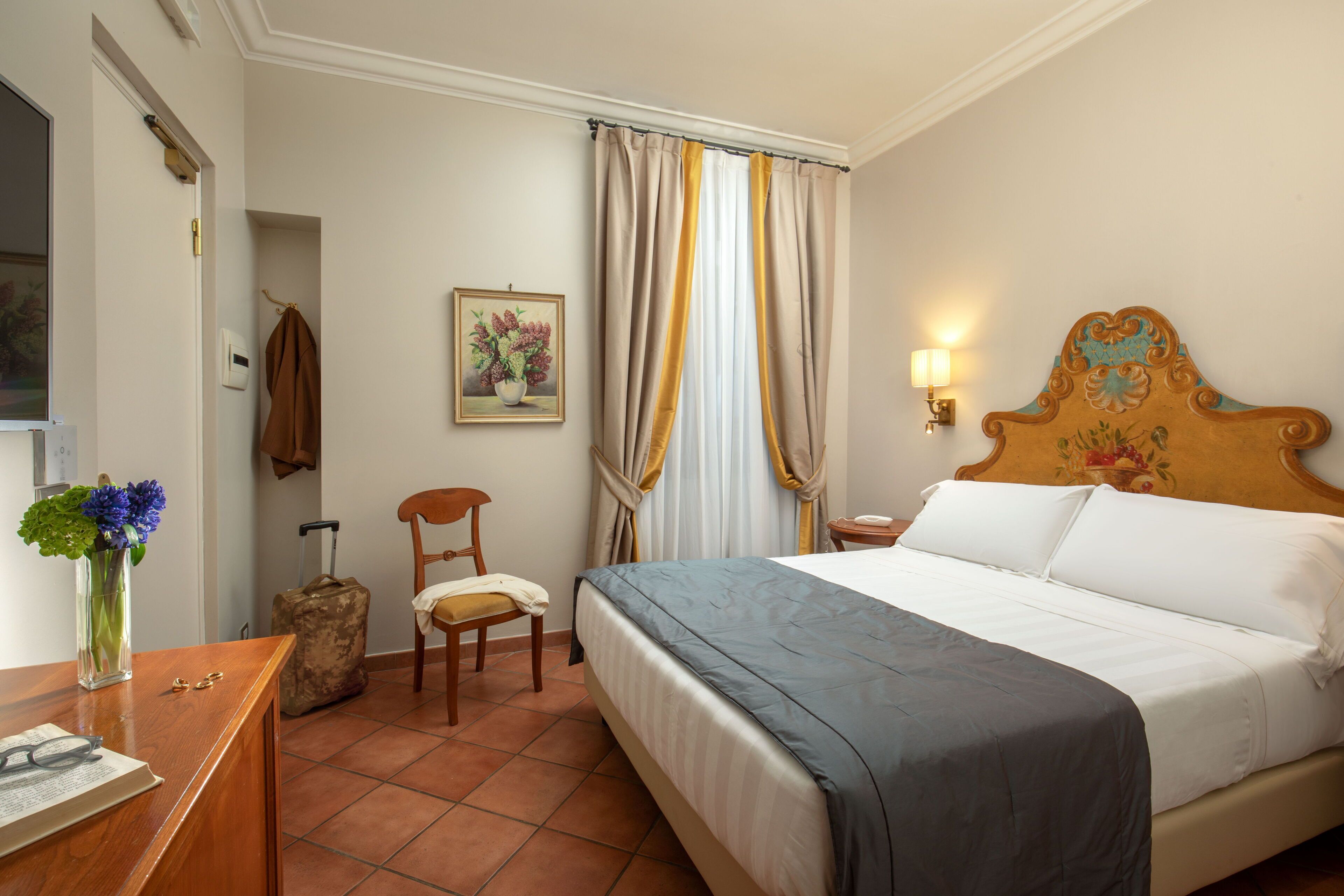 A hotel room with a bed, a chair, a desk, a lamp, a painting, and a suitcase.