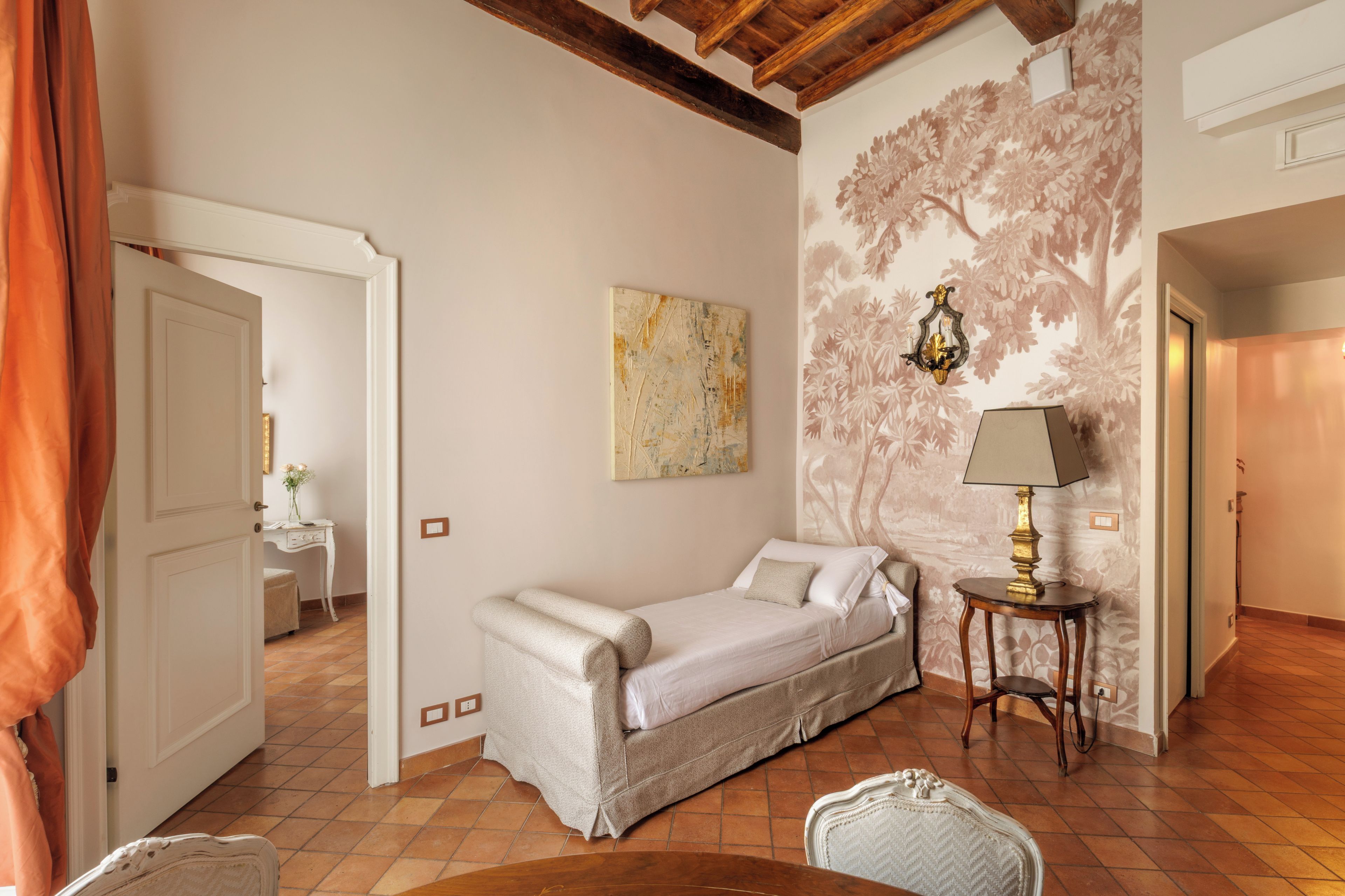 A bedroom with a single bed, a small table with a lamp, a decorative wall, and a door leading to another room.