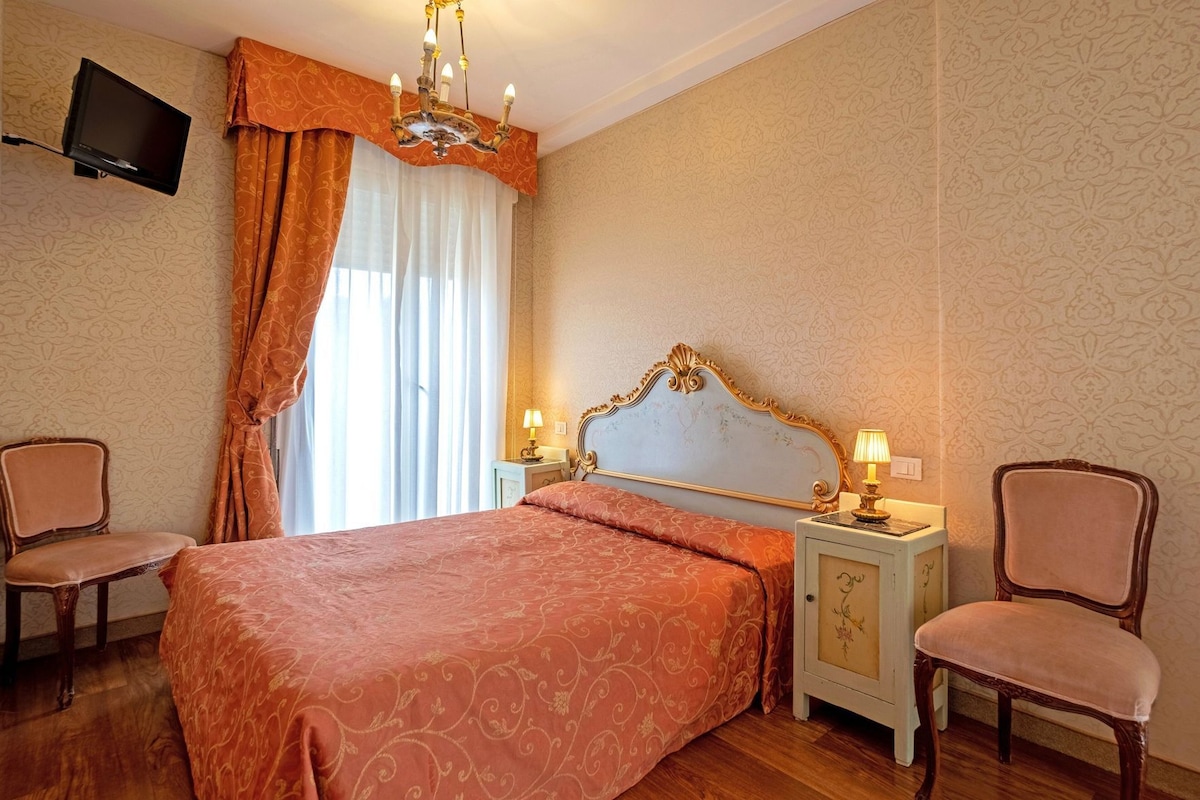A hotel room with a large bed, two bedside tables with lamps, a TV mounted on the wall, and a chandelier.