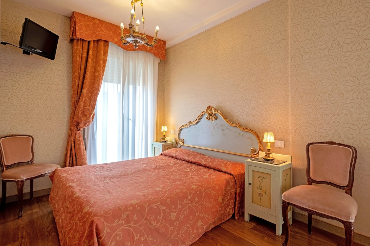 A bedroom with a large bed, two chairs, a nightstand with lamps, a television on the wall, and patterned wallpaper.