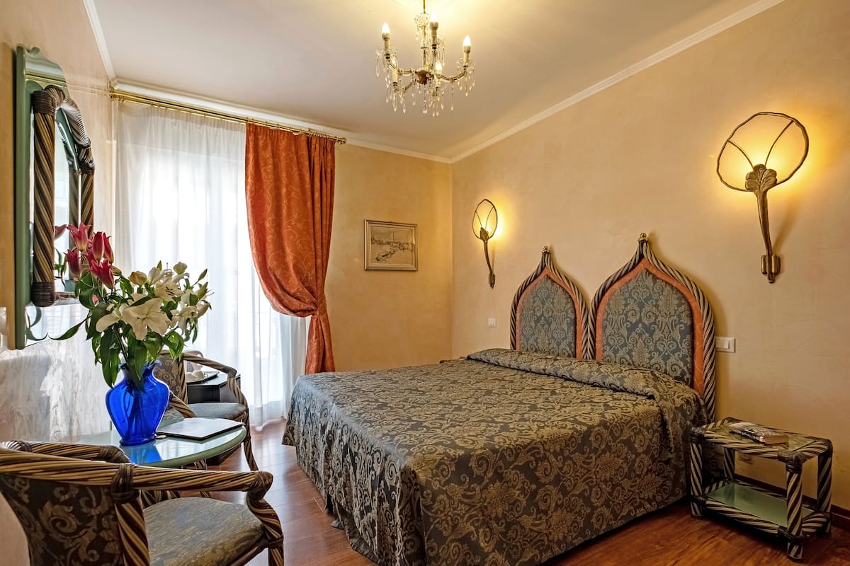 A hotel room with a bed, a chair, a table, a vase of flowers, a chandelier, and wall sconces.
