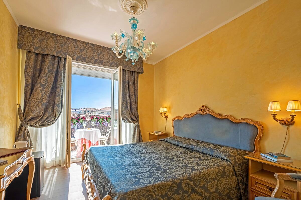 A hotel room with a large bed, a chandelier, a balcony with a table and chairs, and a view of the city.