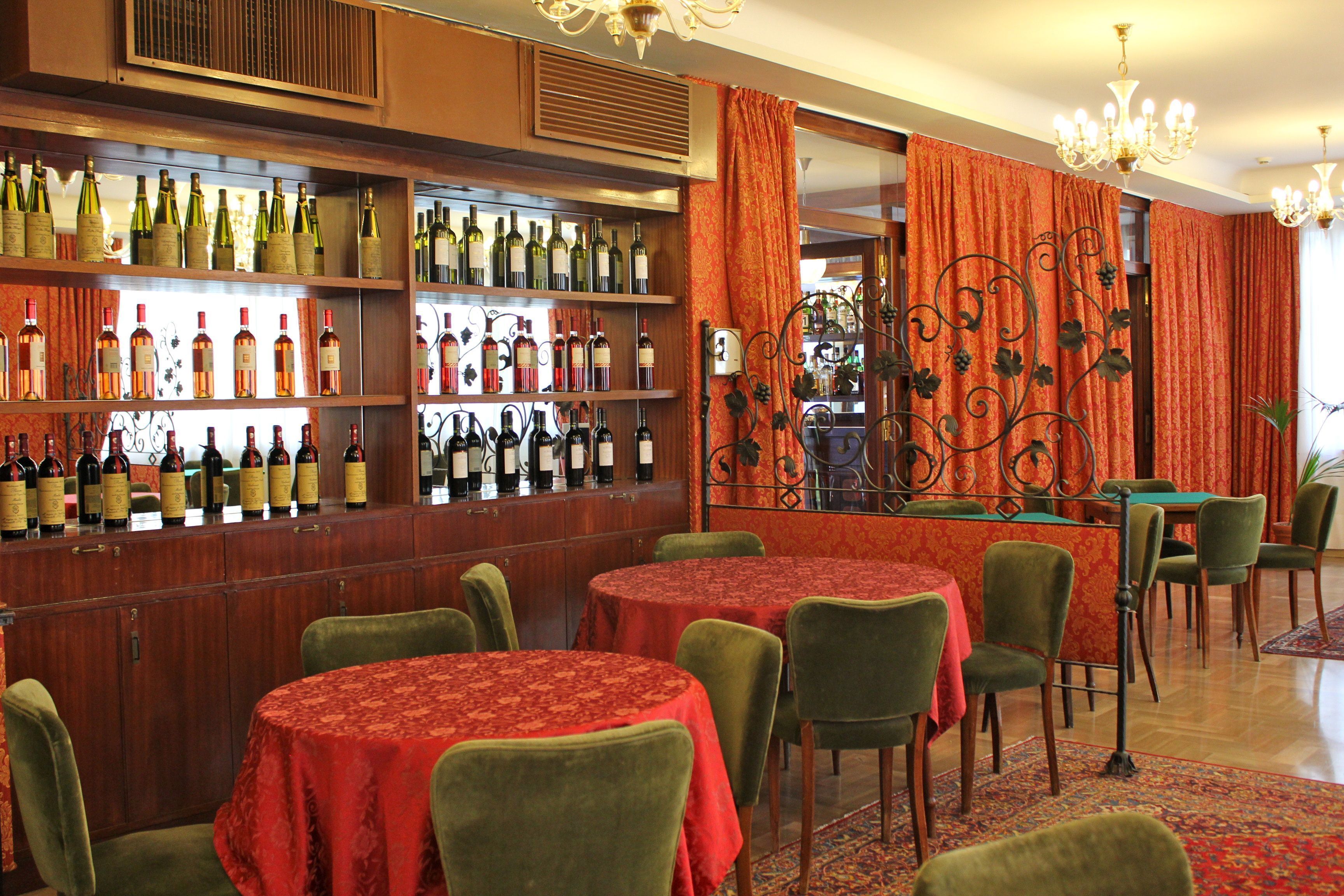 Una zona de bar con múltiples estantes de botellas de vino, mesas redondas con manteles rojos y sillas tapizadas en verde.
