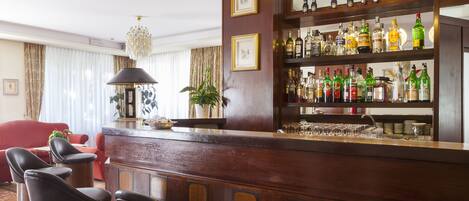 A bar area with a wooden counter, multiple liquor bottles, glasses, and a chandelier. There are three black bar stools and a red sofa in the background.