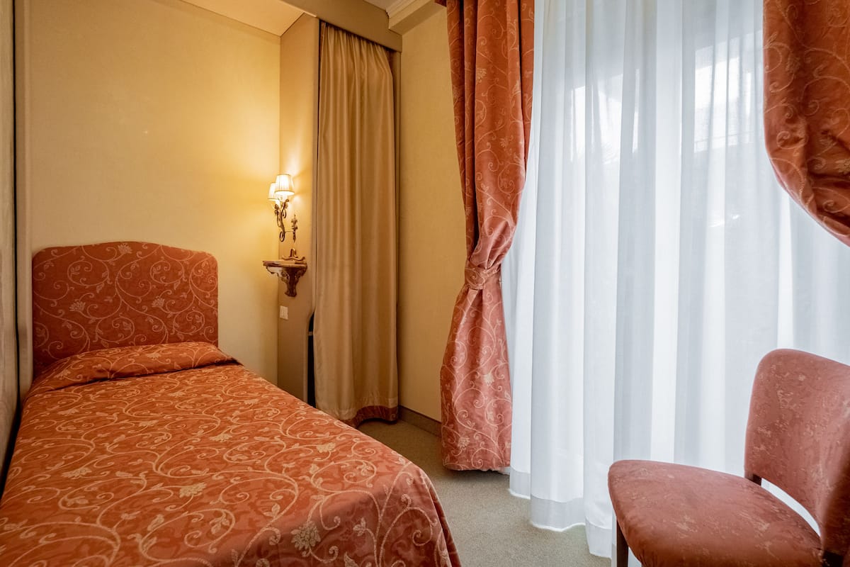 A hotel room with a bed, a chair, a nightstand with a lamp, and sheer curtains.