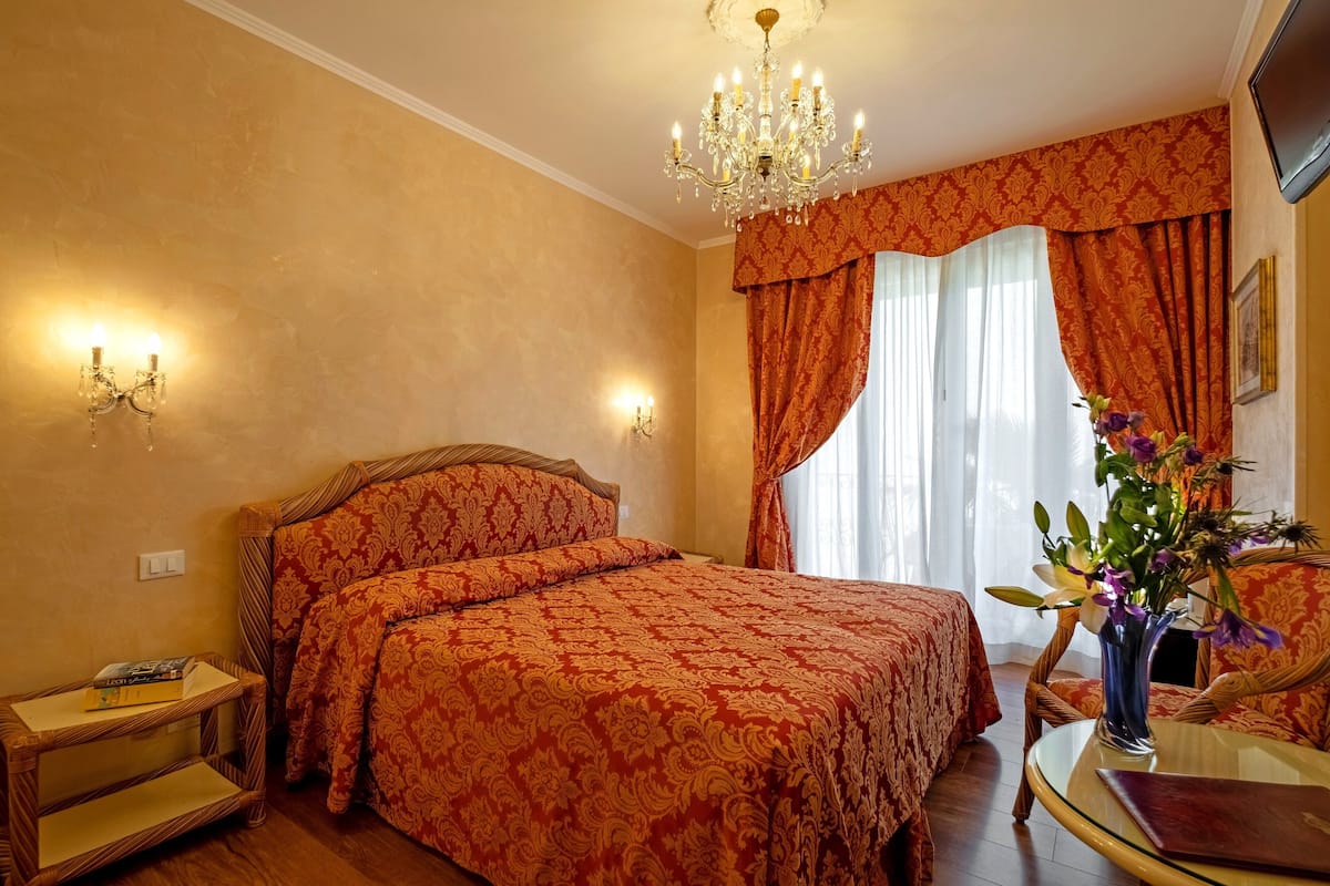 A hotel room with a large bed, a chandelier, a glass table, and a vase with flowers.