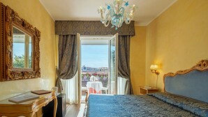Ett hotellrum med en stor säng, ett skrivbord med en stol, en kristallkrona och utsikt över en balkong med blommor.