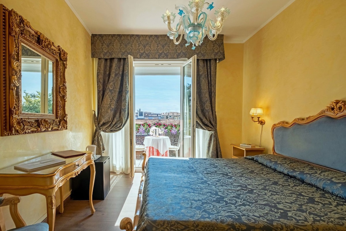 A hotel room with a bed, desk, chair, and a view of the city through the window.