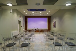 Una sala conferenze con un grande schermo, file di sedie e un podio.