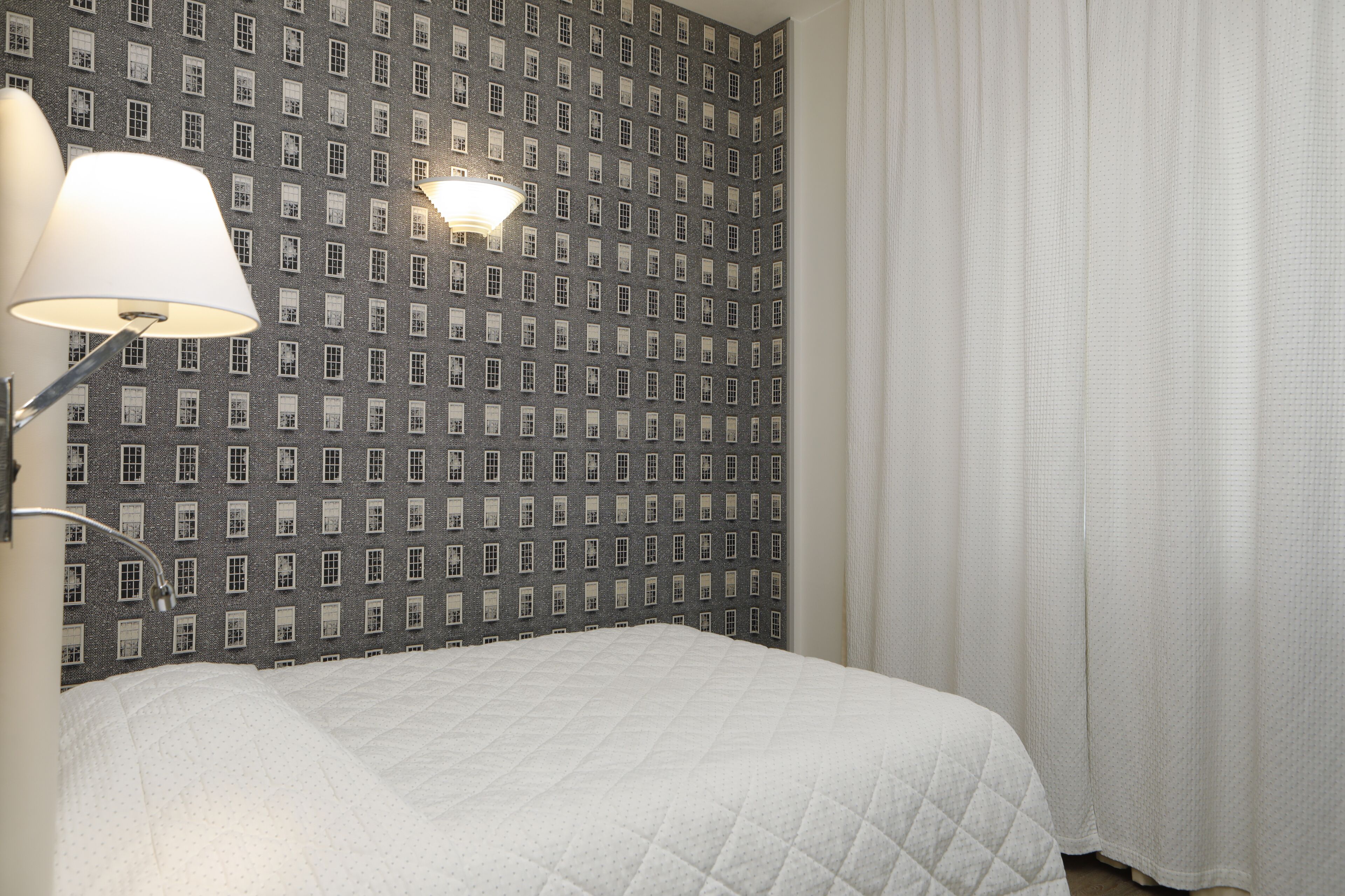 A hotel room with a patterned wallpaper, a bed with a quilt, a lamp, and a window with curtains.