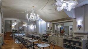 Un comedor amplio con varias mesas y sillas, un candelabro central y una zona de cocina con mesas de buffet.