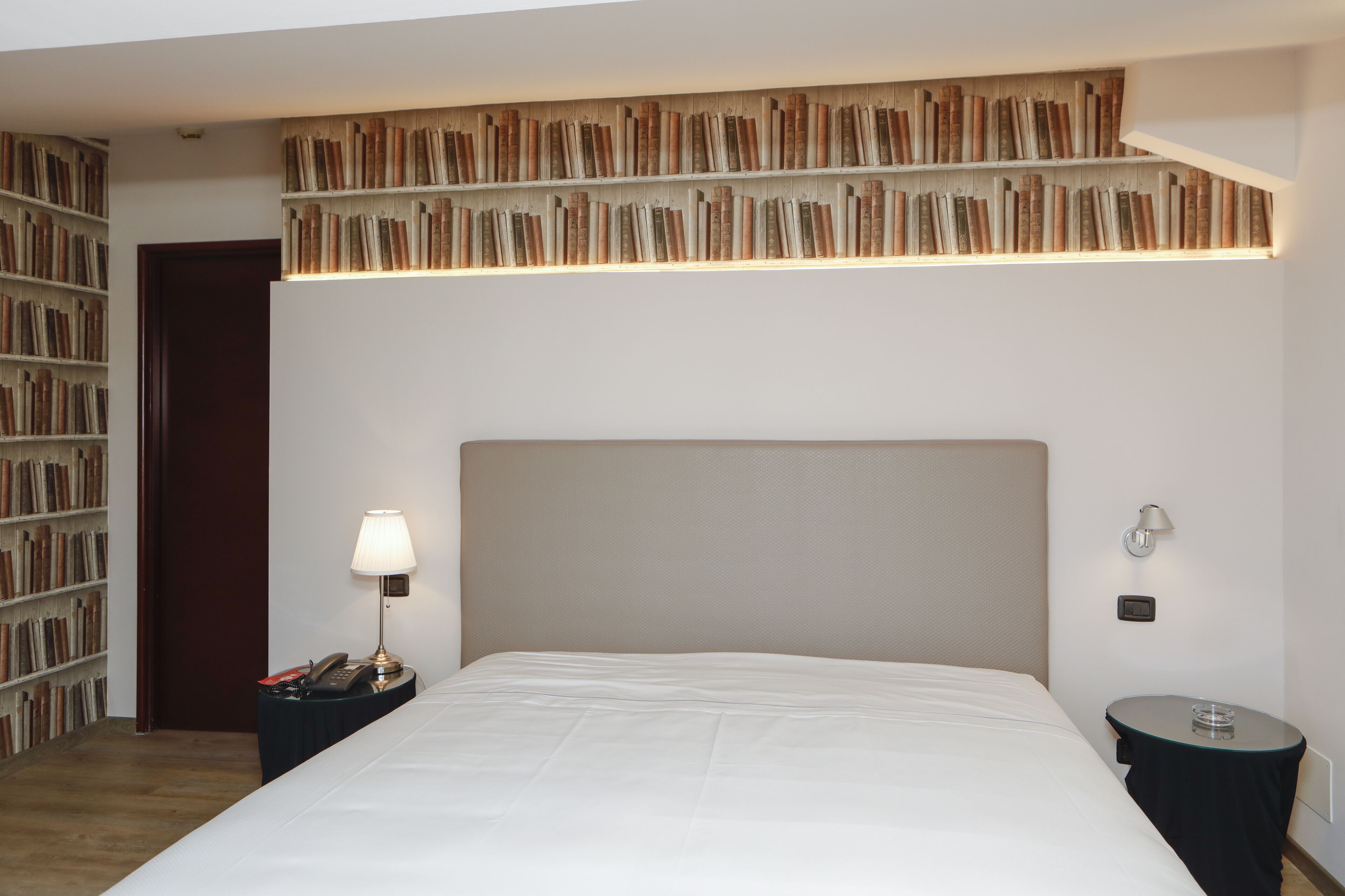 A bedroom with a large bed, a bedside table with a lamp, a small side table, and a bookshelf against the wall.