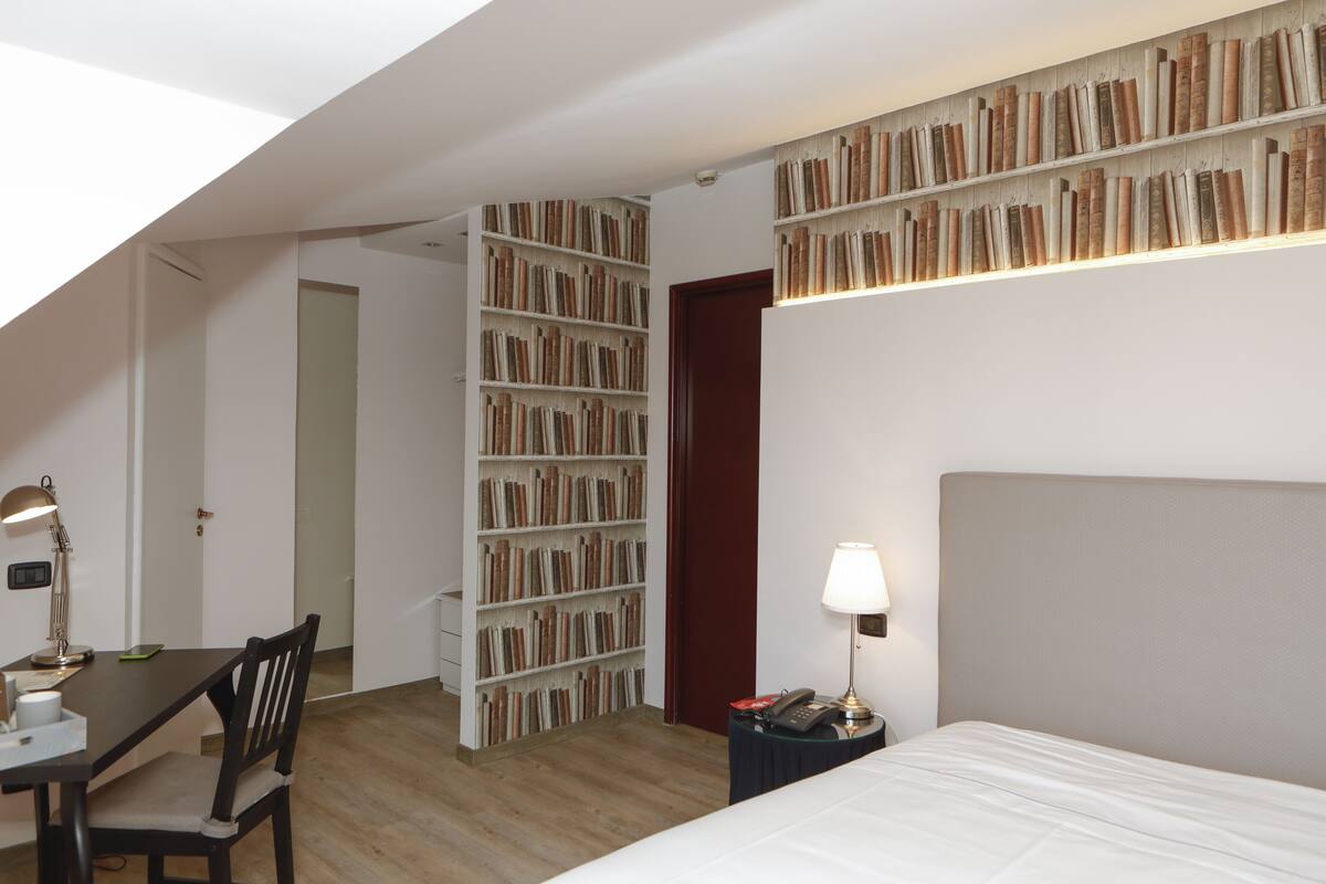 A bedroom with a bed, a desk with a chair, a bookshelf, and a nightstand with a lamp.