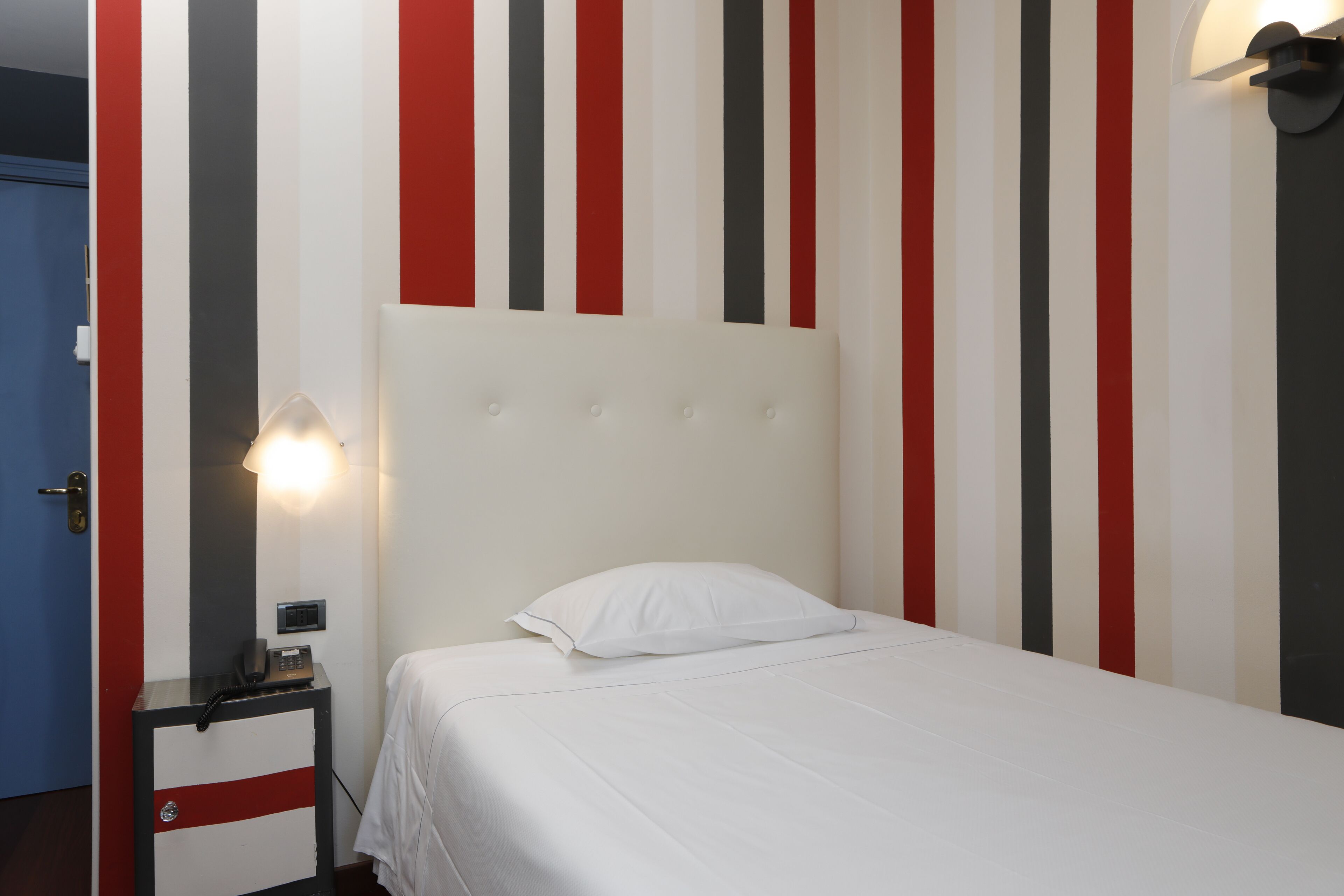Una habitación de hotel con una cama, una mesita de noche con lámpara y una pared a rayas verticales.