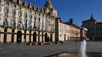 Ein historischer Platz mit einem Brunnen, Gebäuden im klassischen Stil und blauem Himmel.