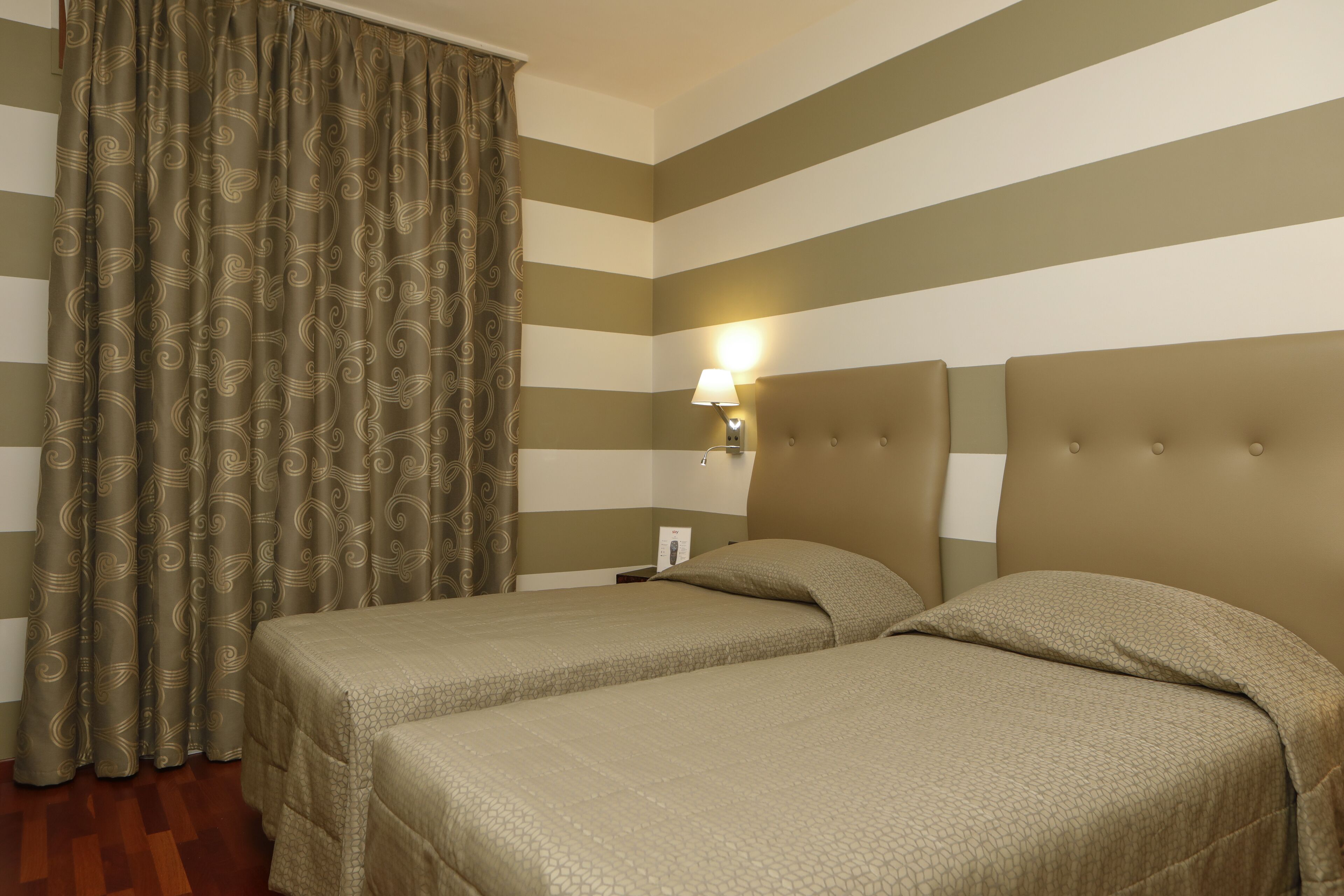 Habitación de hotel con dos camas, paredes a rayas, una cortina y una mesita de noche con lámpara.