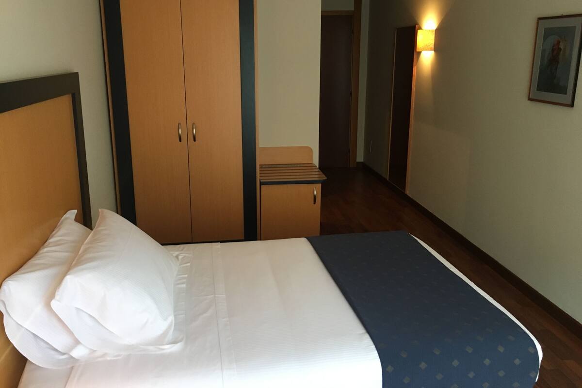 A hotel room with a bed, a wardrobe, a small table, and a hallway with a light.