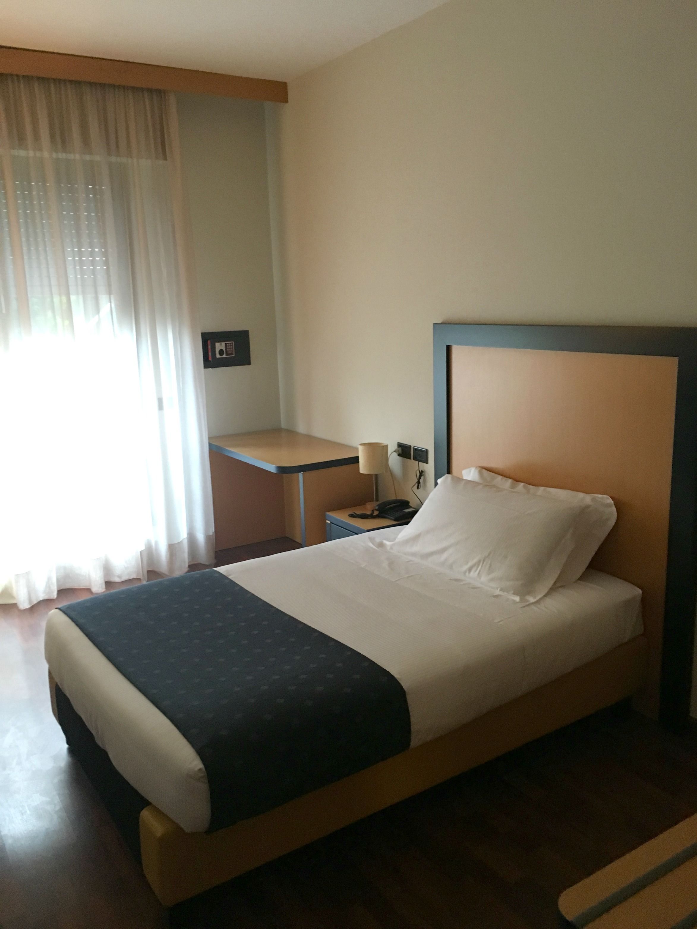 A hotel room with a bed, bedside table, lamp, and a small desk with a telephone.