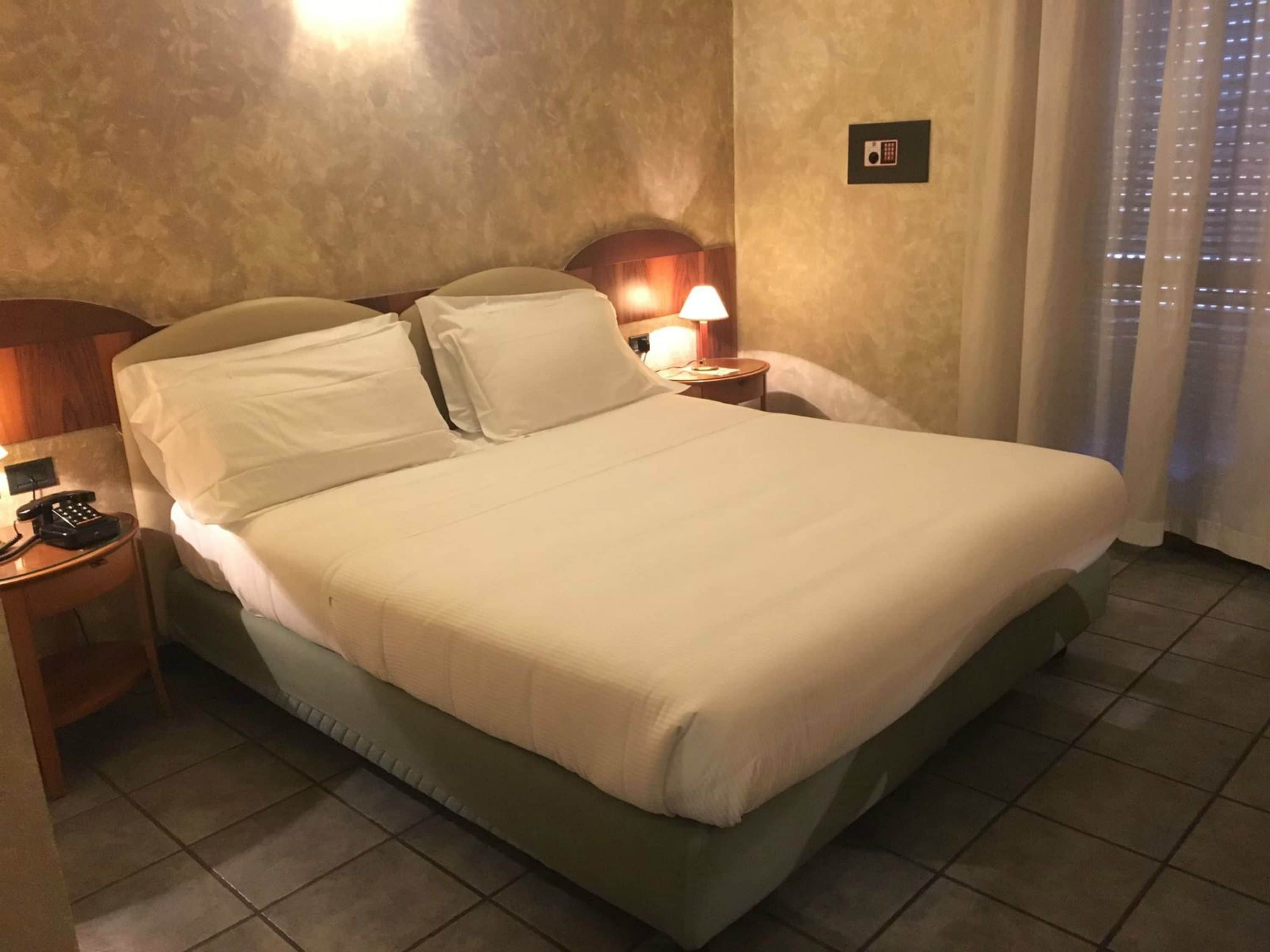 A hotel room with a large bed, a bedside table with a lamp, a phone, and a wall-mounted light switch.