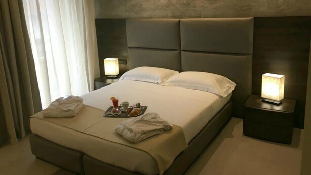 A modern hotel room with a large bed, two bedside tables with lamps, a tray with breakfast items, and a view of the outdoors through sheer curtains. - Plus Welcome Milano (Milan)